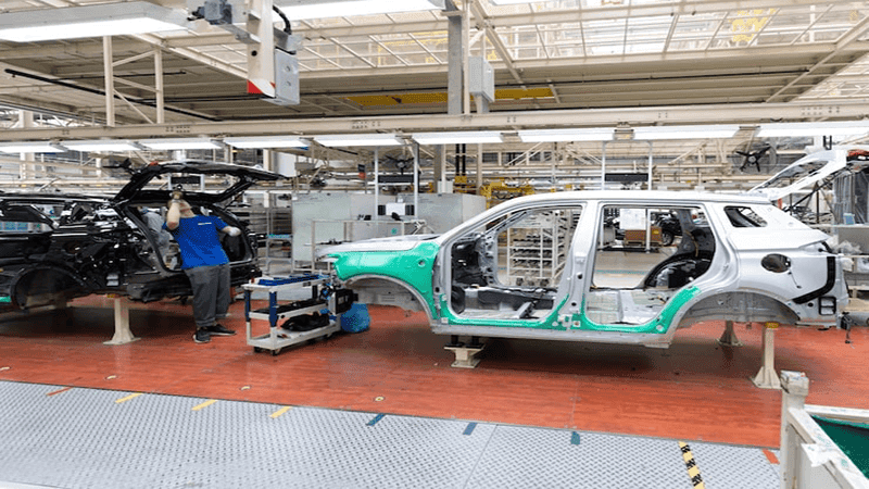 Automotive assembly line: Factory worker in blue uniform assembling car body frames (white and black) on a manufacturing plant floor during vehicle production