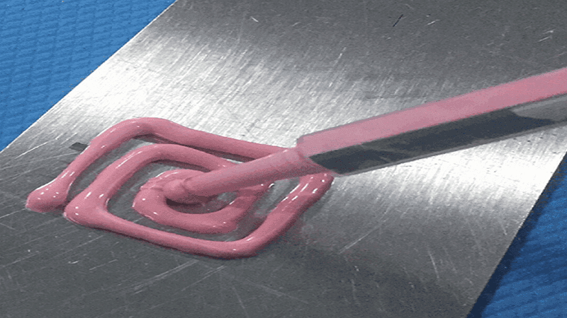 Pink adhesive sealant application: Syringe dispensing viscous pink adhesive in a spiral pattern onto a brushed metal sheet, used for industrial bonding, sealing, or assembly processes