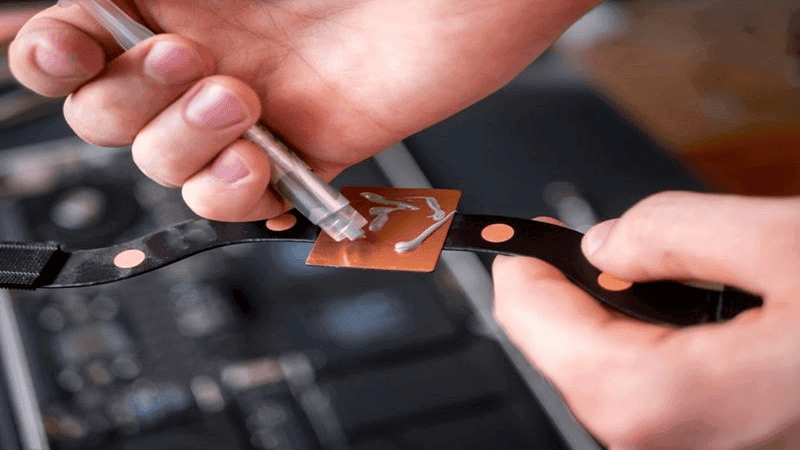 Solder paste application on flexible PCB: Hands dispensing solder paste onto a copper pad on a black flexible circuit board, a key step in electronics assembly and printed circuit board (PCB) manufacturing