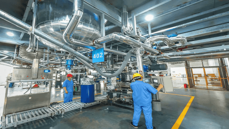 Industrial manufacturing facility: Factory workers in safety helmets (red, yellow) operating equipment near large stainless steel piping, tanks, and machinery on a plant floor for process industry operations