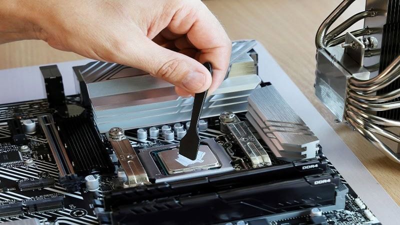 A hand using a small black spatula to spread white thermal paste evenly across the silver surface of a CPU (Central Processing Unit) installed on a motherboard, with a large CPU cooler visible on the right.